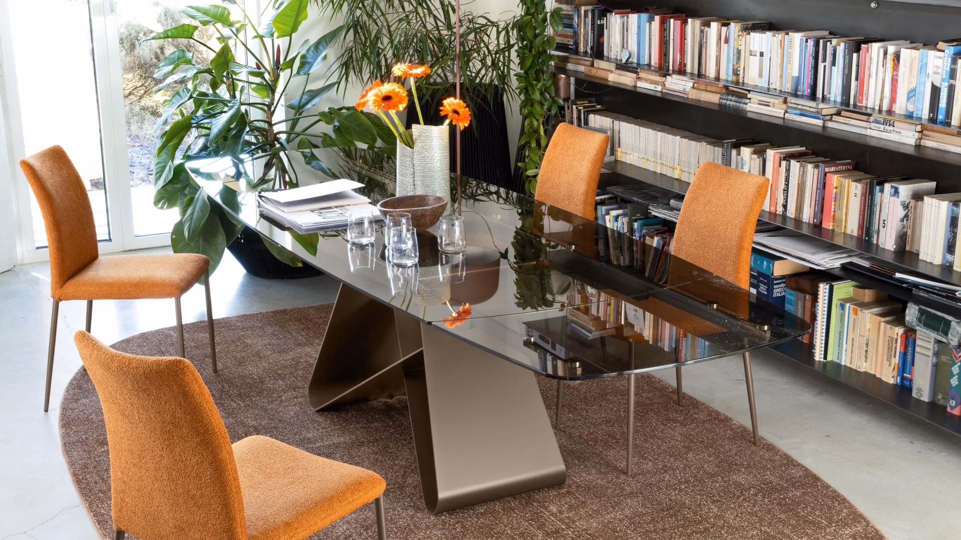 Connubia orange upholstered dining chairs around a glass table beside bookshelves and indoor plants.