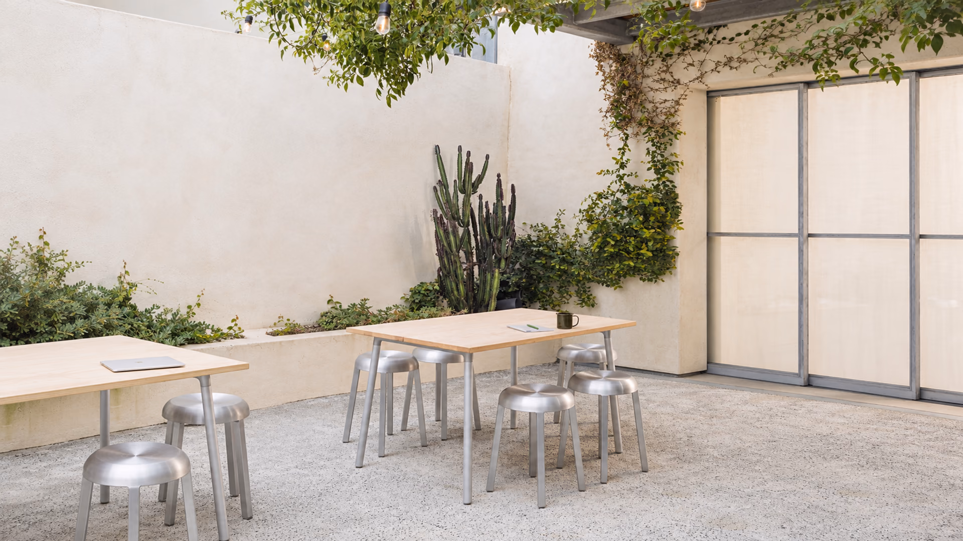 Emeco brushed aluminum stools and light wood tables in a courtyard with climbing greenery.