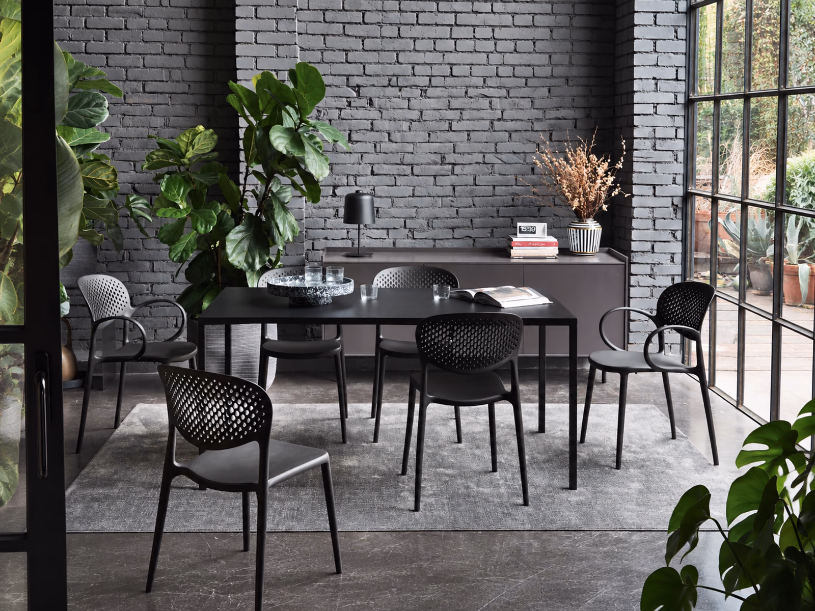 Connubia: An Iron table and perforated chairs form a dining setting against dark brick and tall windows.