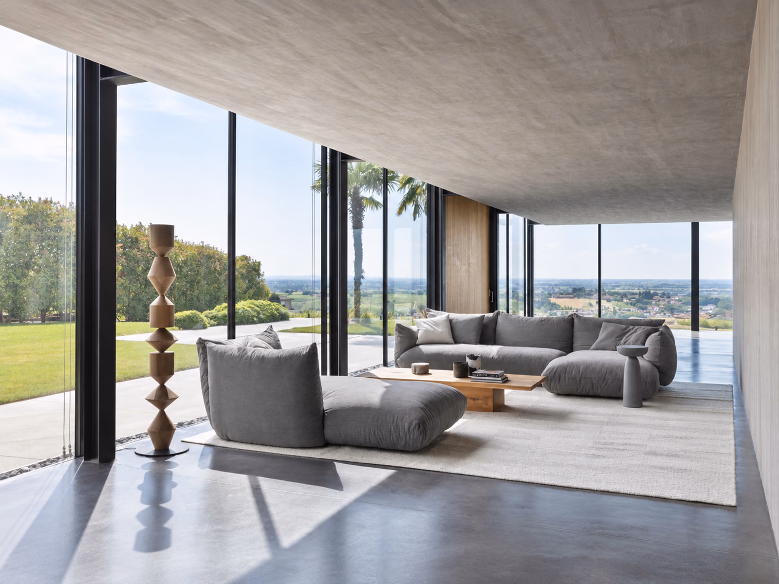 Mogg: Gray Alba sofa seating surrounds a wood coffee table in a glass-walled room overlooking the landscape.