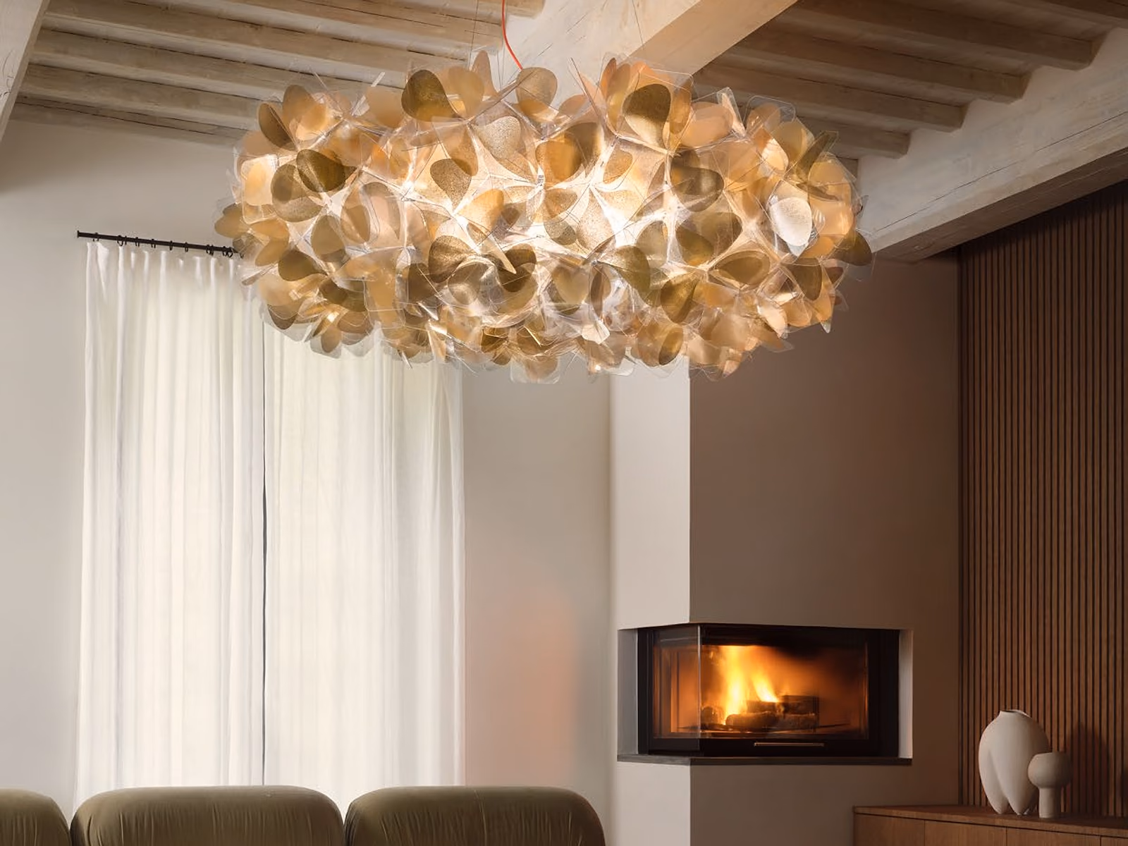 Slamp: A gold Clizia pendant glows above low olive sofas beside a corner fireplace.