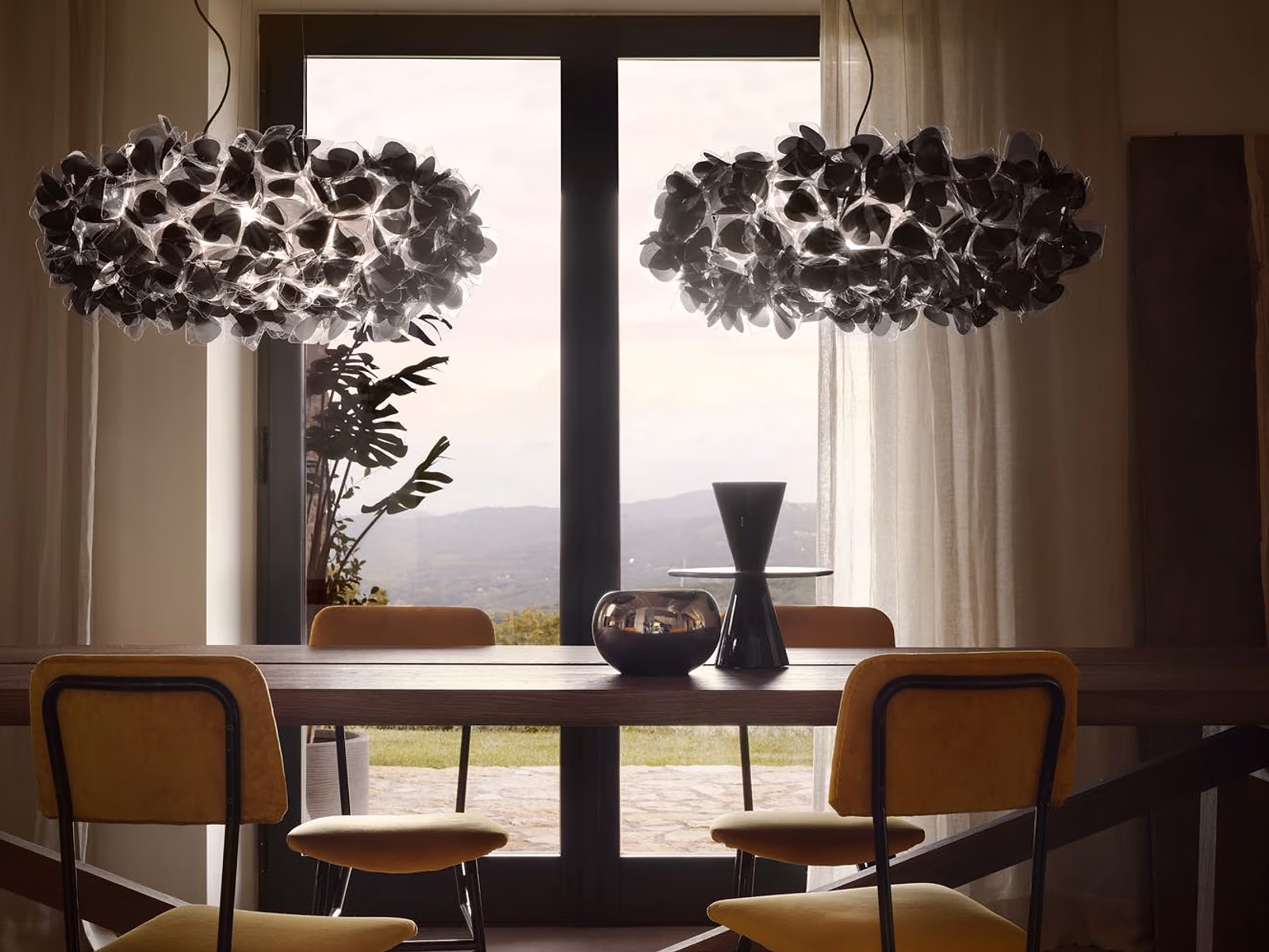 Slamp: Two smoked Clizia pendants hang above a wood dining table in front of a wide garden window.